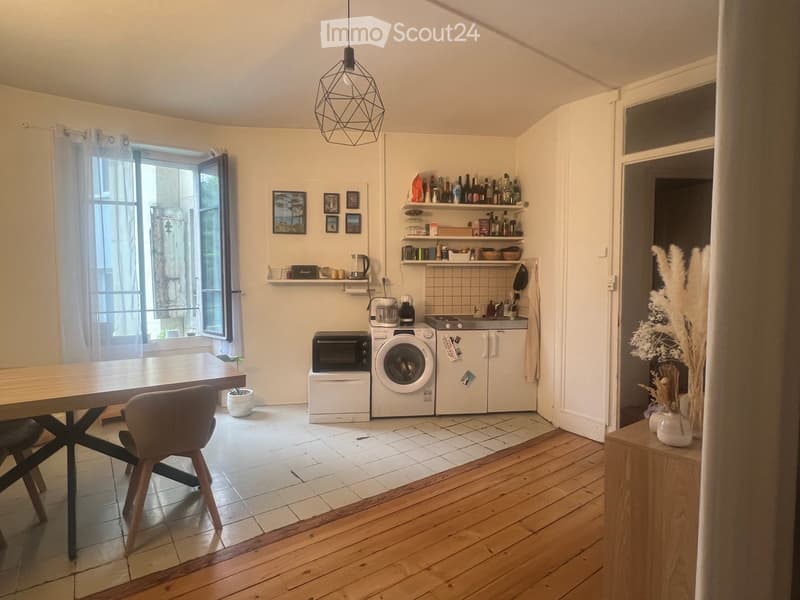 Bright kitchen with wooden flooring, white tiled backsplash, and modern appliances including a microwave, washing machine, and refrigerator. Features a wooden dining table with chairs, shelves with bottles and kitchen items, and a pendant light hanging fr