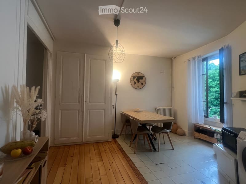 Bright room with wooden floor, large window, dining table with chairs, vase with dried plants, radiator, washing machine, and bench.