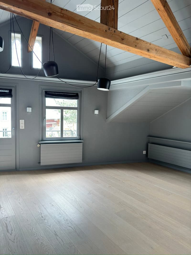Empty attic room with wooden beams, exposed ceiling, wooden flooring, and large windows. No furniture or equipment present.
