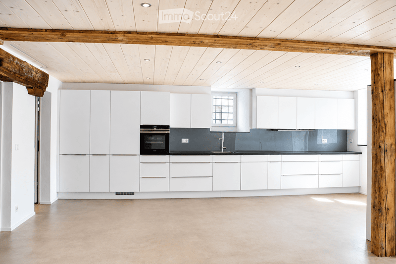Modern kitchen with white cabinets, a built-in oven, a sink, and a faucet. The space features a light-colored concrete floor and exposed wooden beams on the ceiling.