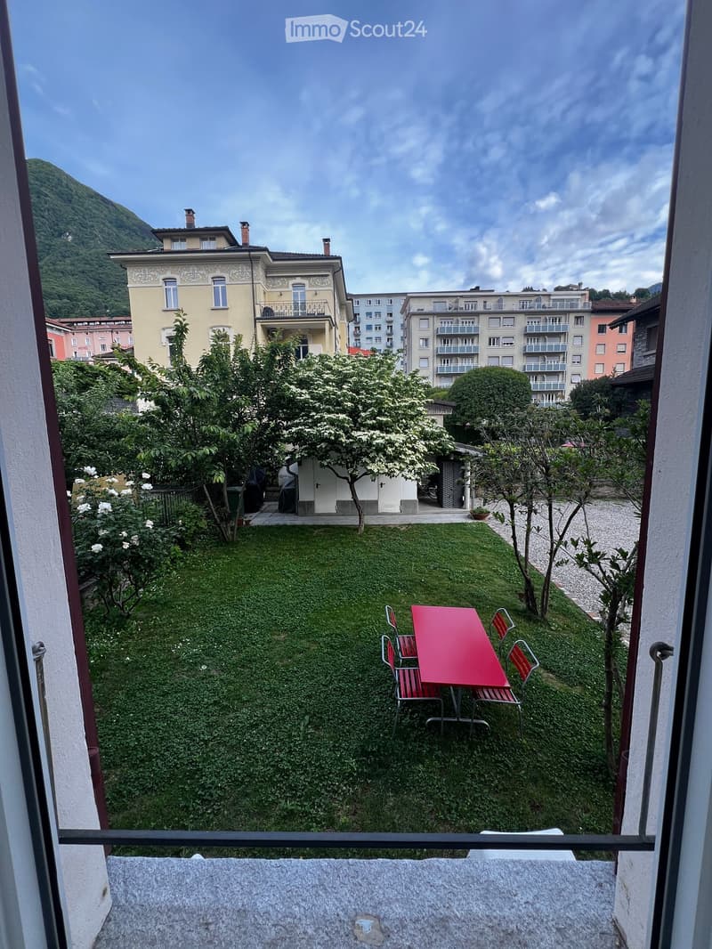 Garden area with a red table and chairs, surrounded by greenery and trees, overlooking a cityscape with buildings and a mountain in the background.