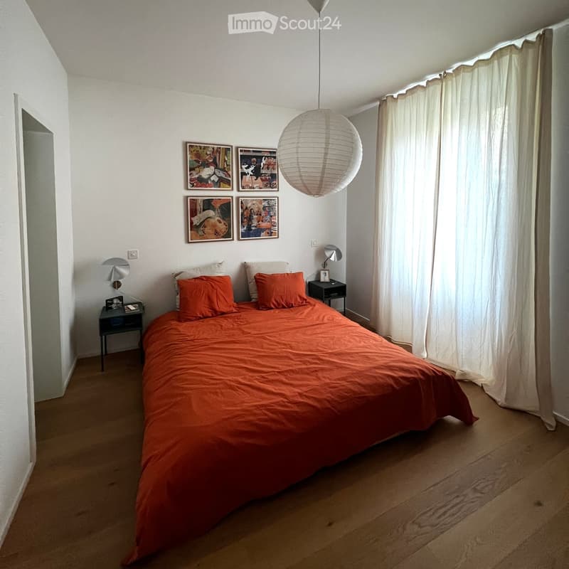 A cozy bedroom with a single bed covered in an orange duvet, two nightstands with lamps, and several picture frames on the wall. The room has wooden flooring and white walls.