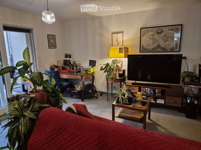 A cozy living room with a red sofa, coffee table, TV, and several potted plants. A balcony door is visible on the left side.