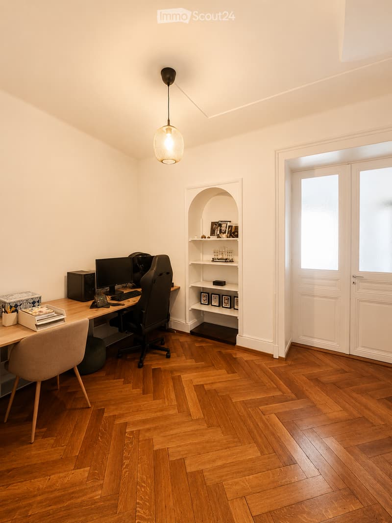 A modern, well-lit study or home office with wooden herringbone flooring, a large wooden desk, ergonomic chair, computer setup, and built-in shelving with decorative items and framed photos.