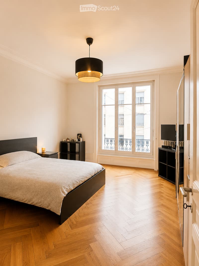 Modern bedroom with wooden herringbone flooring, large window with balcony view, black bed frame, nightstand, TV stand, and pendant light fixture.