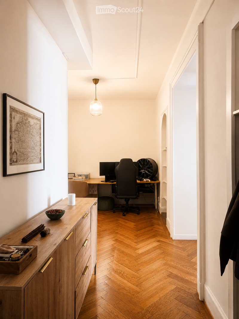 A modern, well-lit room with herringbone wooden flooring, a desk with a computer and chair, a hanging lamp, a wooden cabinet with drawers, and decorative items on the cabinet.