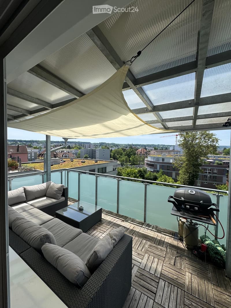 Modern balcony with wooden decking, glass railing, grey wicker sofa, coffee table, and grill.