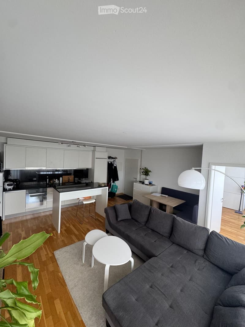 Modern open-plan living and dining area with a grey sectional sofa, white coffee table, white stools, a kitchen island with bar stools, and a dining table with chairs. The space features light grey walls, wooden flooring, and contemporary furniture.