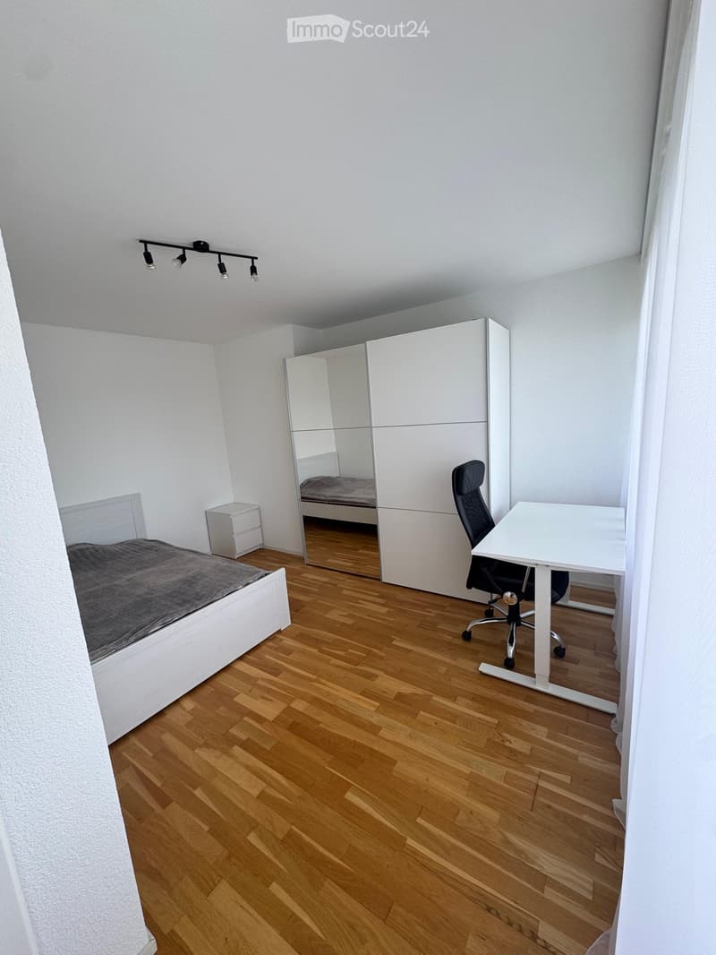 A modern, well-lit bedroom with a wooden floor, a white bed with a grey blanket, a white wardrobe with a mirrored door, a desk with a chair, and a small nightstand.