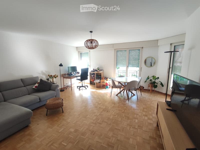 Bright living room with wooden flooring, large windows, and a balcony. Features a grey sofa, a round dining table with chairs, a TV screen, and a desk with a computer.