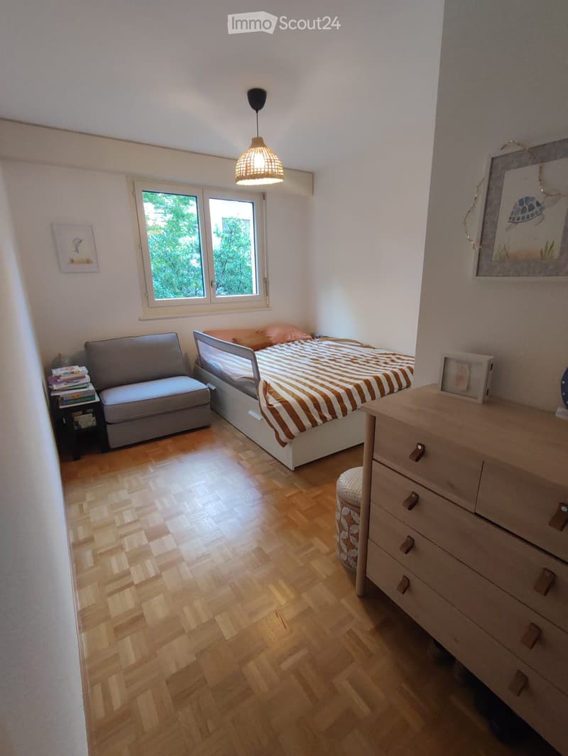 Cozy bedroom with wooden flooring, a bed with striped bedding, a sofa, a nightstand, a drawer, a small stool, and a hanging lamp.