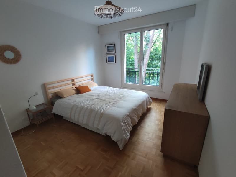 Bright bedroom with wooden flooring, a double bed with a white duvet and orange pillows, a nightstand with a lamp, a wardrobe, and a large window with a view of greenery.