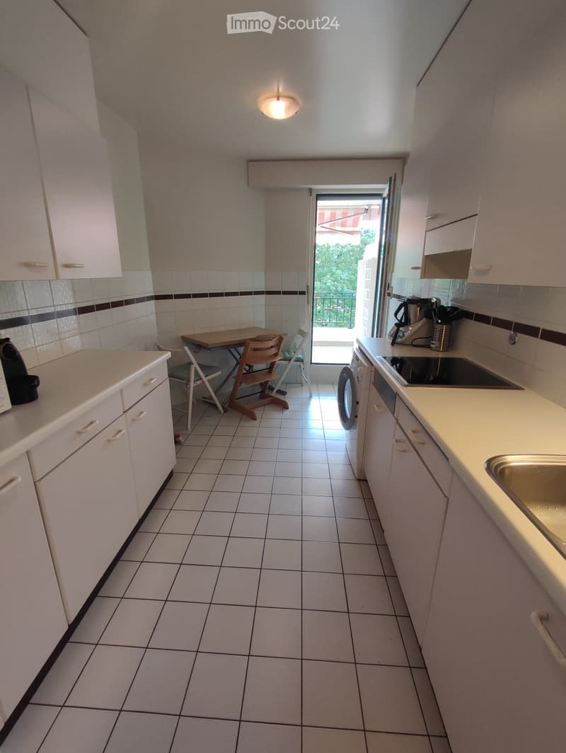 A small, well-lit kitchen with white cabinets, tiled floors, and a small dining area. The kitchen features a sink, fridge, oven, coffee maker, and a small table with chairs.