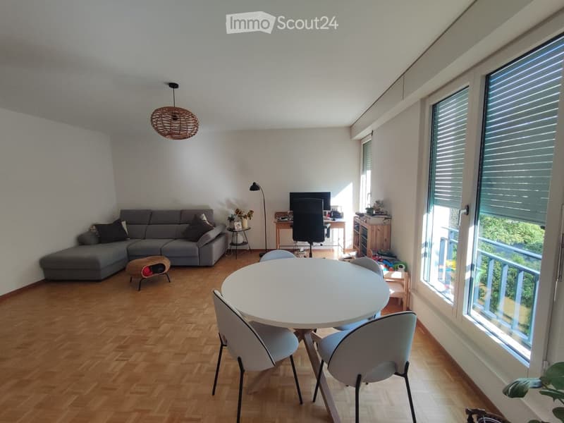 A modern living room with a grey sectional sofa, a round white dining table with four chairs, a coffee table, a hanging wicker lamp, and a TV screen. Large windows with blinds offer a view of greenery outside.