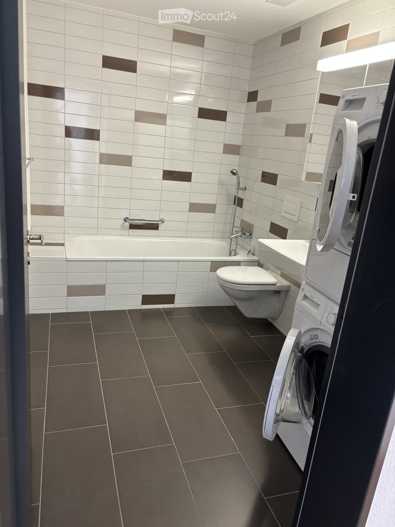 Modern bathroom with white and brown tiled walls, a bathtub, toilet, sink, and a washing machine in the corner.