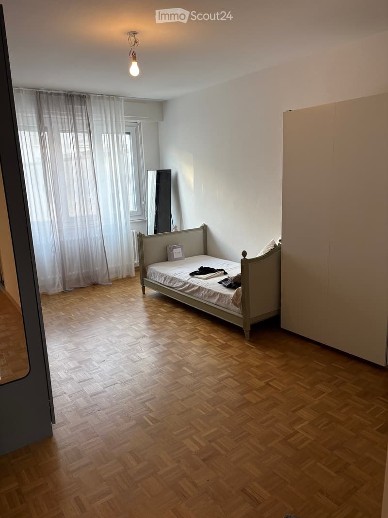 Bedroom with wooden herringbone flooring, a single bed with a grey headboard, a wardrobe, and a mirror. Light-colored curtains cover the window, and the room is minimally furnished.