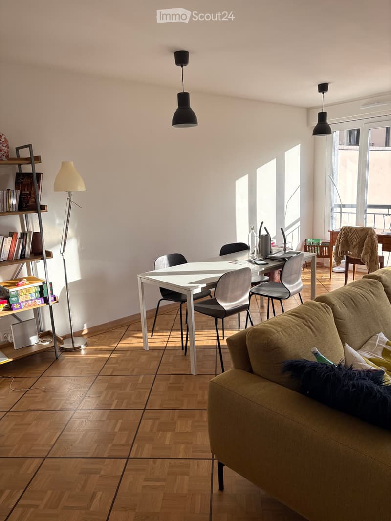 A modern living room with a sofa, coffee table, dining table, chairs, bookshelf, and floor lamp. The room features wooden flooring and large windows allowing natural light.