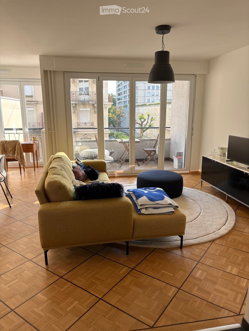 Living room with wooden flooring, large windows leading to a balcony, sofa, ottoman, coffee table, and TV cabinet.