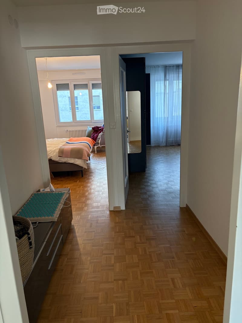 Hallway with wooden flooring leading to a bedroom and another room, featuring a bed, chair, and window in the adjacent bedroom.