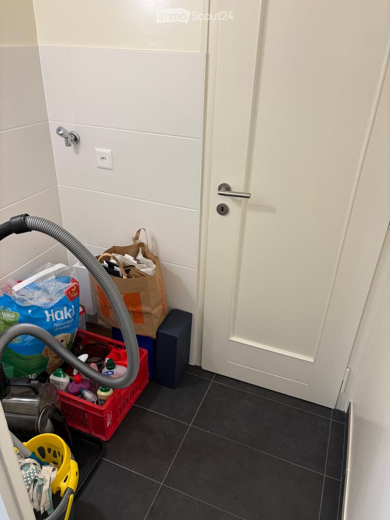 A narrow hallway with a closed white door, tiled walls, and a vacuum cleaner hose extending from the left side. Cleaning supplies and bags are placed on the floor near the door.