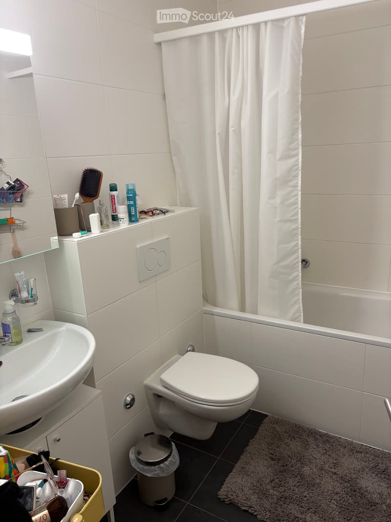 Modern bathroom with white tiles, a sink, toilet, bathtub with a shower curtain, and various toiletries on the counter.