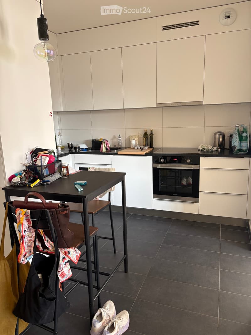 Modern kitchen with white cabinets, black countertop, and stainless steel appliances. Includes a small table with a chair, various kitchen items, and personal belongings.