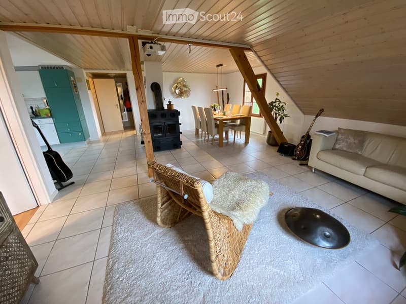Open-plan living and dining area with a sofa, wicker chair, dining table, and fireplace. The space features wooden beams, tiled flooring, and a cozy rug.