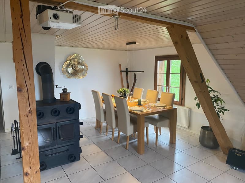 Cozy dining area with a wooden table, six chairs, a black wood-burning stove, and a projector mounted on the ceiling. The room features exposed wooden beams, large windows, and decorative elements including a wall-mounted floral design and a coffee grinde