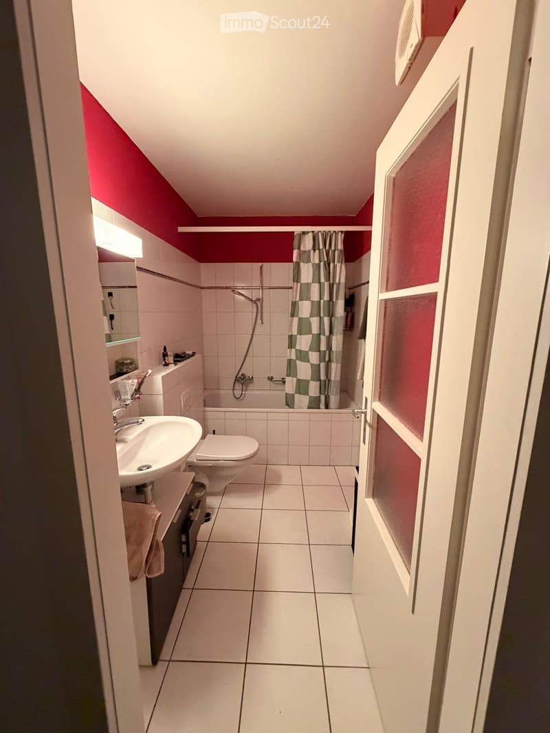 Modern bathroom with white tiles, a sink, toilet, bathtub, and a shower curtain. Features a red accent wall and built-in storage.