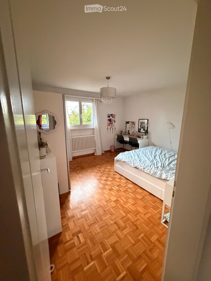 Cozy bedroom with wooden herringbone flooring, white walls, a single bed with light blue bedding, a desk with a chair, a mirror, and a window with white curtains.