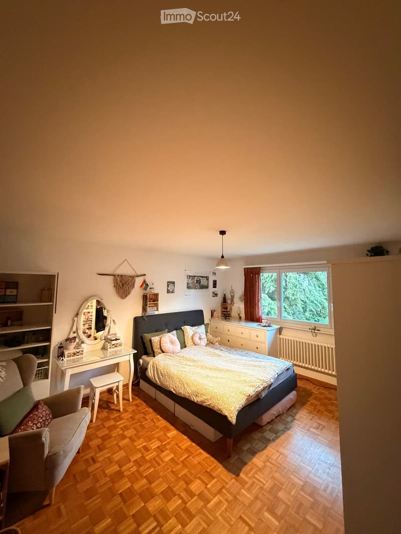 Cozy bedroom with wooden flooring, a large bed with pillows and a patterned bedspread, a vanity table with a mirror, a small chair, and a window with curtains.