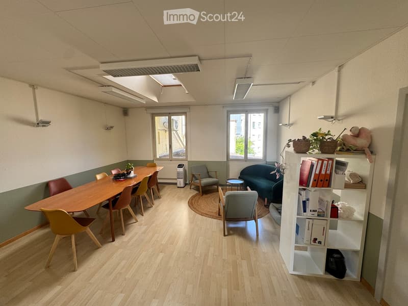 Bright, modern room with wooden flooring, large windows, a long wooden table surrounded by chairs, a grey sofa, a small round stool, a white shelving unit with books and decorative items, and a small plant on the table.