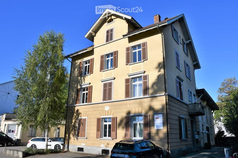 A three-story residential building with beige walls, multiple windows with brown shutters, and a small balcony on the right side. The building is surrounded by trees and parked cars.