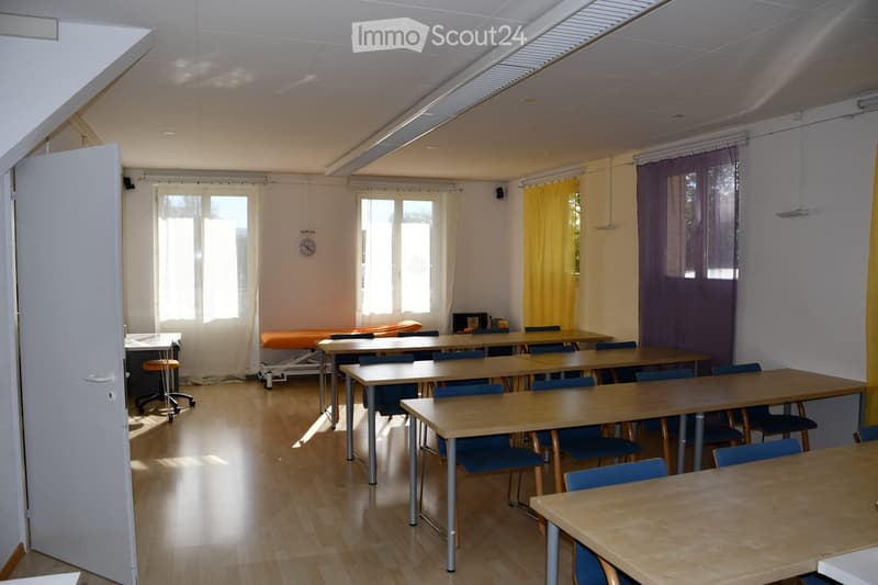 Brightly lit room with wooden flooring, multiple tables and chairs arranged in rows, a small stool, and curtains on the windows.