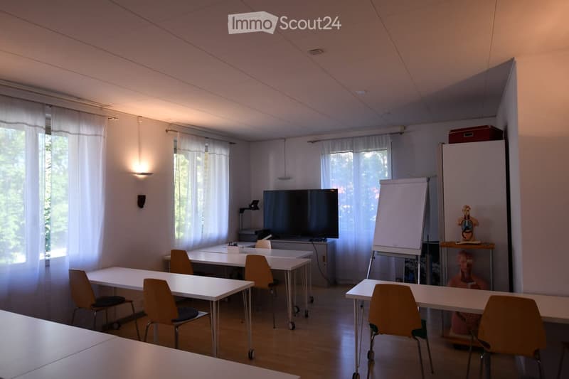 Bright, modern room with white tables, orange chairs, a large screen, whiteboard, and a model on a shelf.