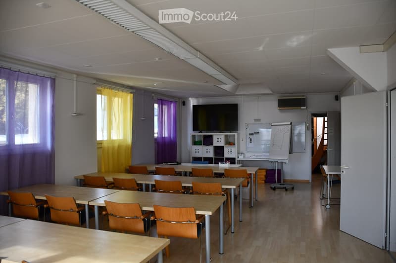 Classroom with multiple tables and orange chairs arranged in rows, a large screen mounted on the wall, a whiteboard, and a small cabinet with storage compartments.