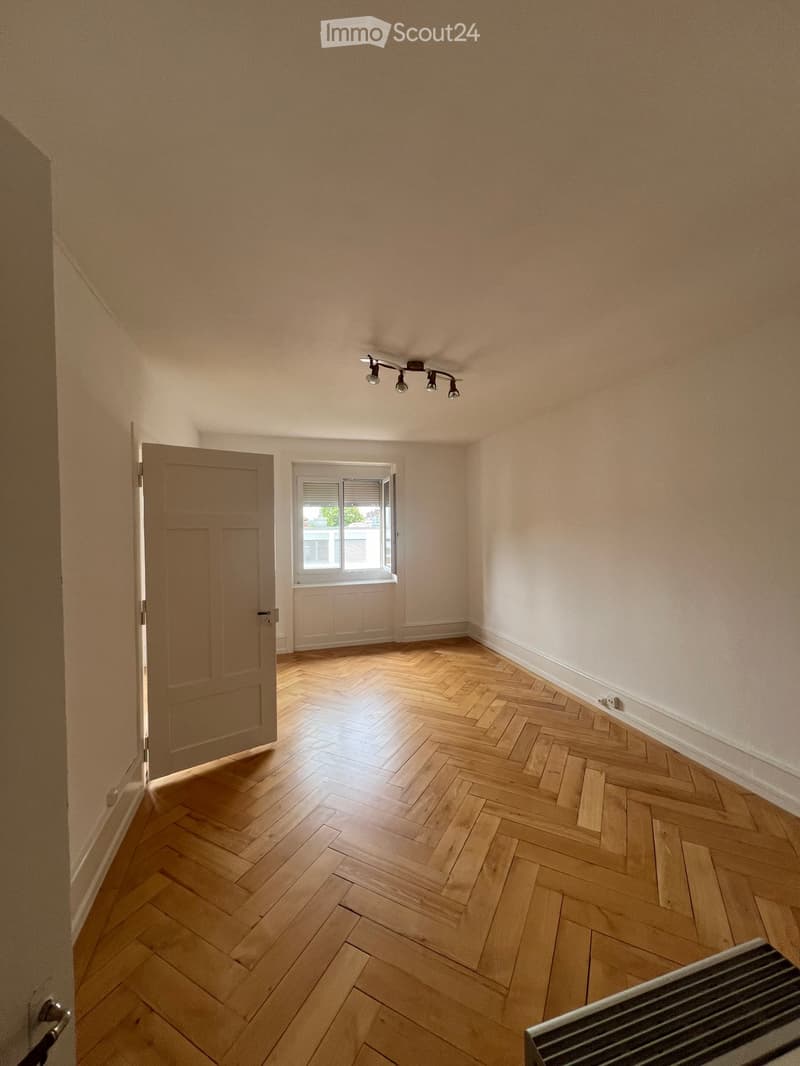 Empty room with wooden herringbone flooring, light-colored walls, a ceiling light fixture, a window with blinds, and a partially visible radiator.