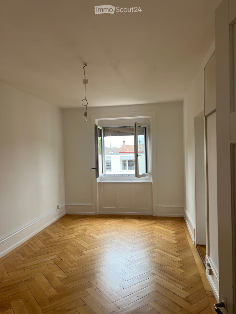 Empty room with wooden herringbone flooring, white walls, and a large window with blinds partially open, allowing natural light to enter.