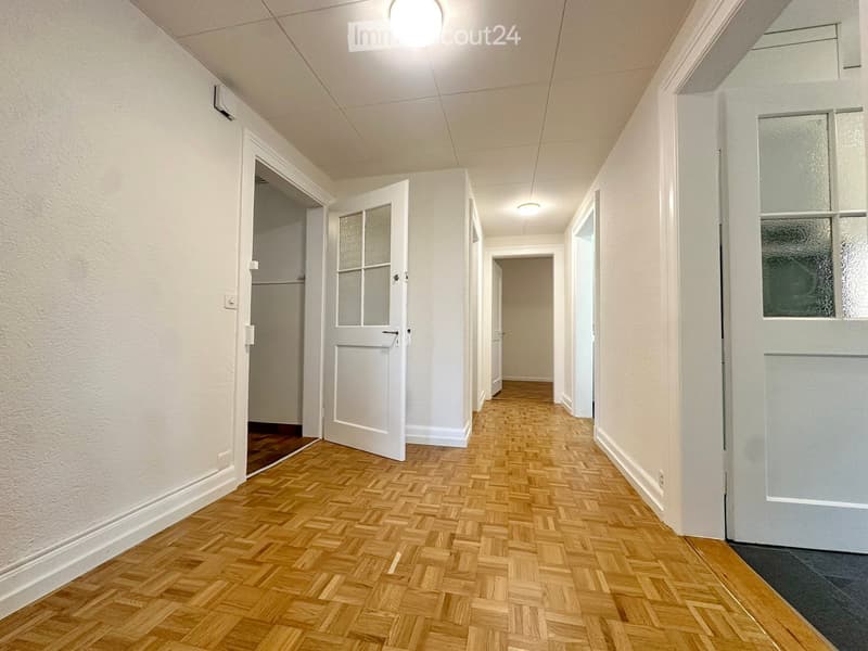 A long, narrow hallway with wooden herringbone flooring, white walls, and several doors with frosted glass panels. The ceiling has recessed lighting and visible air conditioning vents.