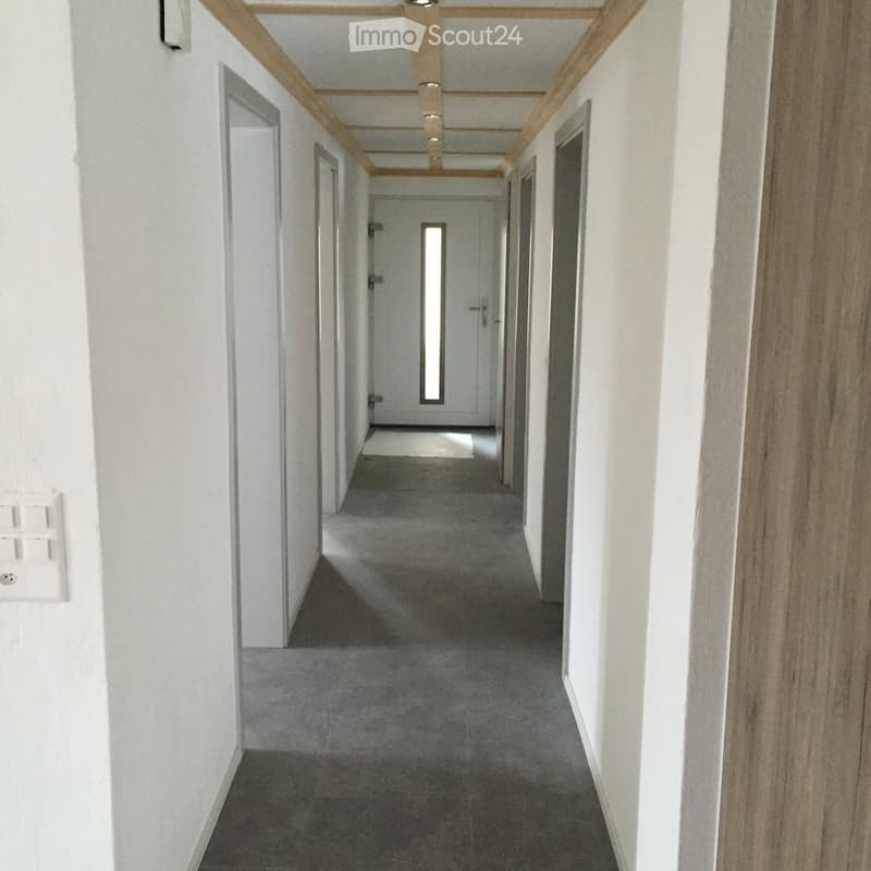 Clean, modern hallway with white walls, grey carpeted floor, and wooden ceiling beams. Doors line both sides, with a small rug placed near the far end.