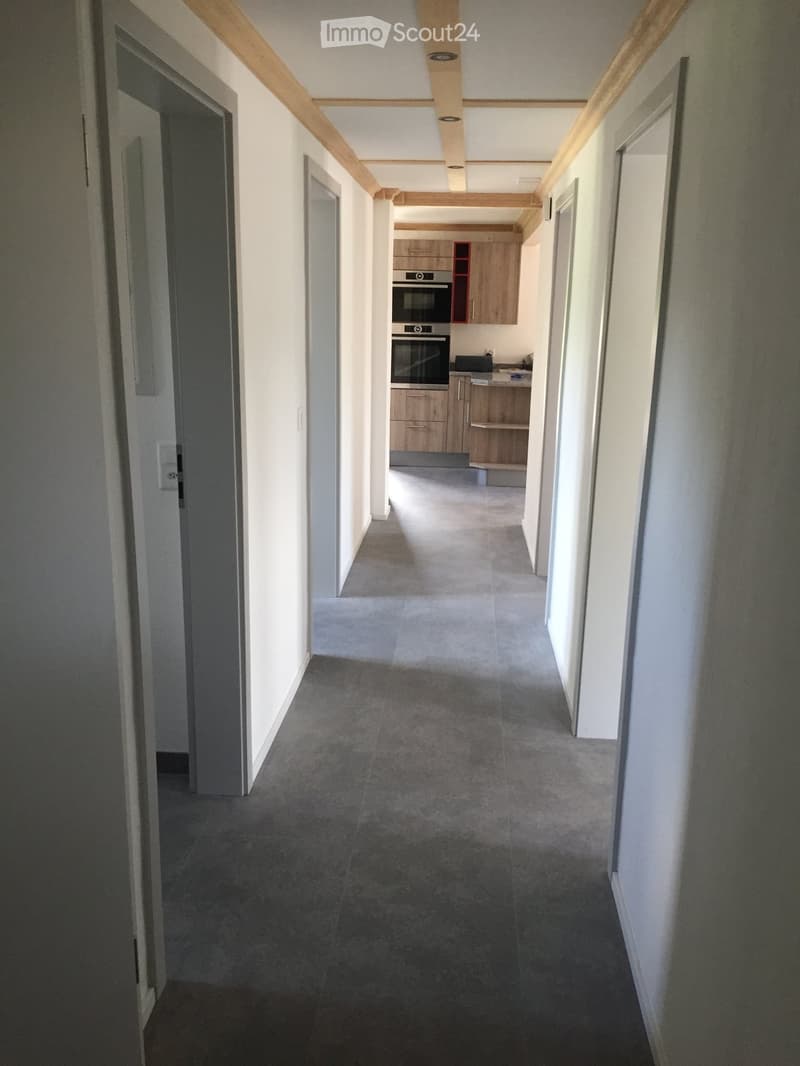 A long, narrow hallway with light-colored walls, dark tiled flooring, and wooden ceiling beams. The hallway leads to a kitchen area with cabinets and appliances visible at the end.