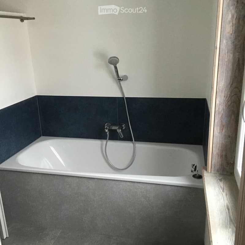Modern bathroom with a white bathtub, dark grey tiles on the walls, and a grey tiled floor. Features a handheld showerhead and a faucet above the tub.