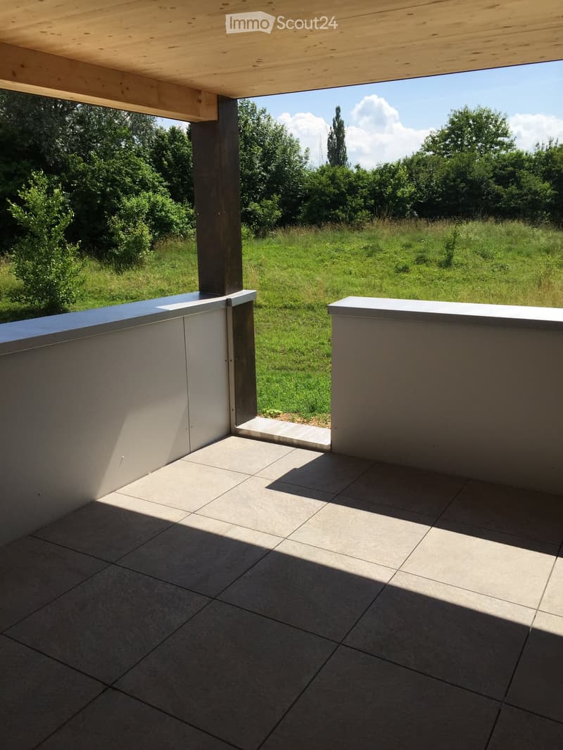 Modern outdoor patio with tiled flooring, wooden pergola, and low white walls, overlooking a grassy field with trees.