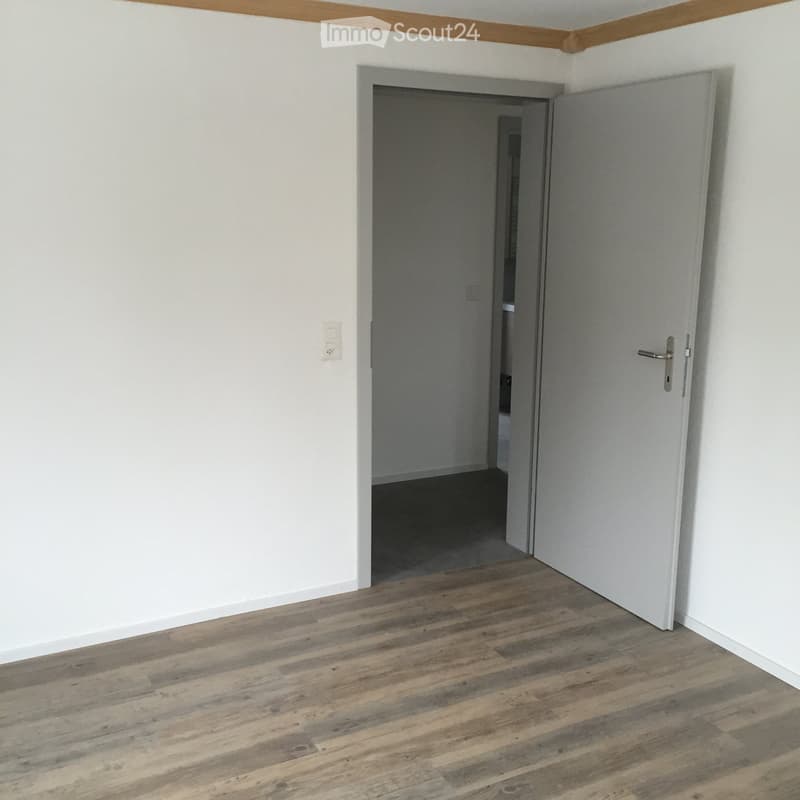 Empty room with light wooden flooring, white walls, and an open door leading to another room.