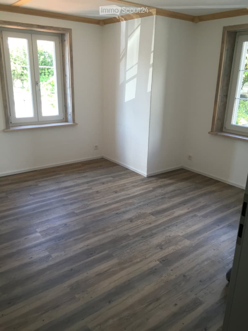 Empty room with wooden flooring, white walls, and two windows with white frames. The room has a simple, clean design with no furniture or decorations.