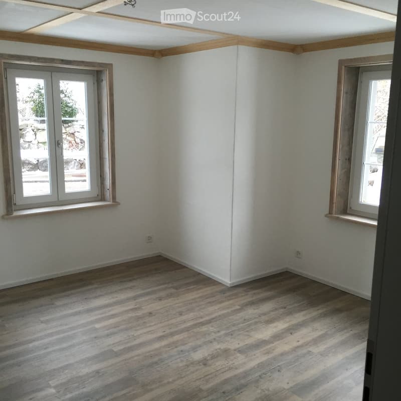 Empty room with light-colored wooden flooring, white walls, and two windows with wooden frames. The ceiling features exposed wooden beams.