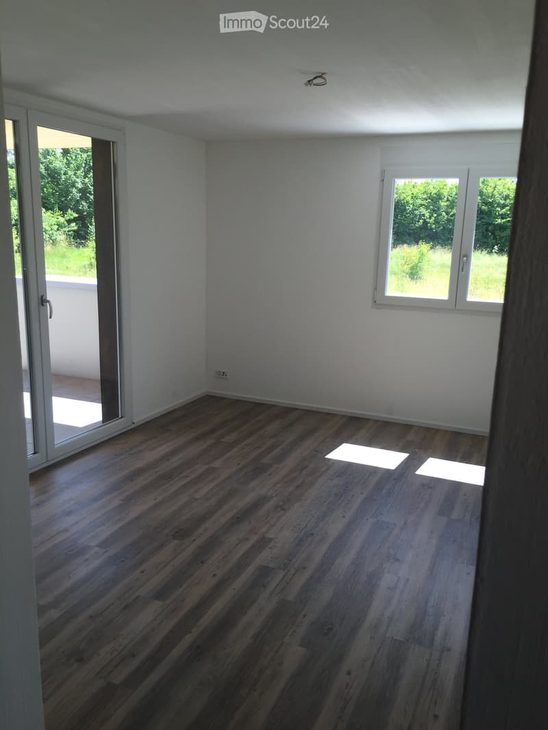 Empty room with wooden flooring, white walls, large windows, and a sliding glass door leading to a balcony.