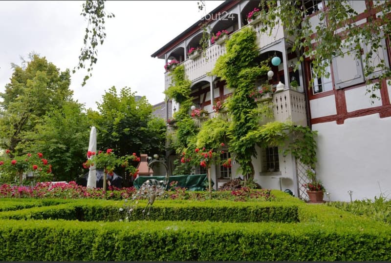 A well-maintained garden with neatly trimmed hedges, colorful flowers, and a decorative sculpture. A charming, multi-story house with white walls, red trim, and balconies adorned with plants and flowers is visible in the background.