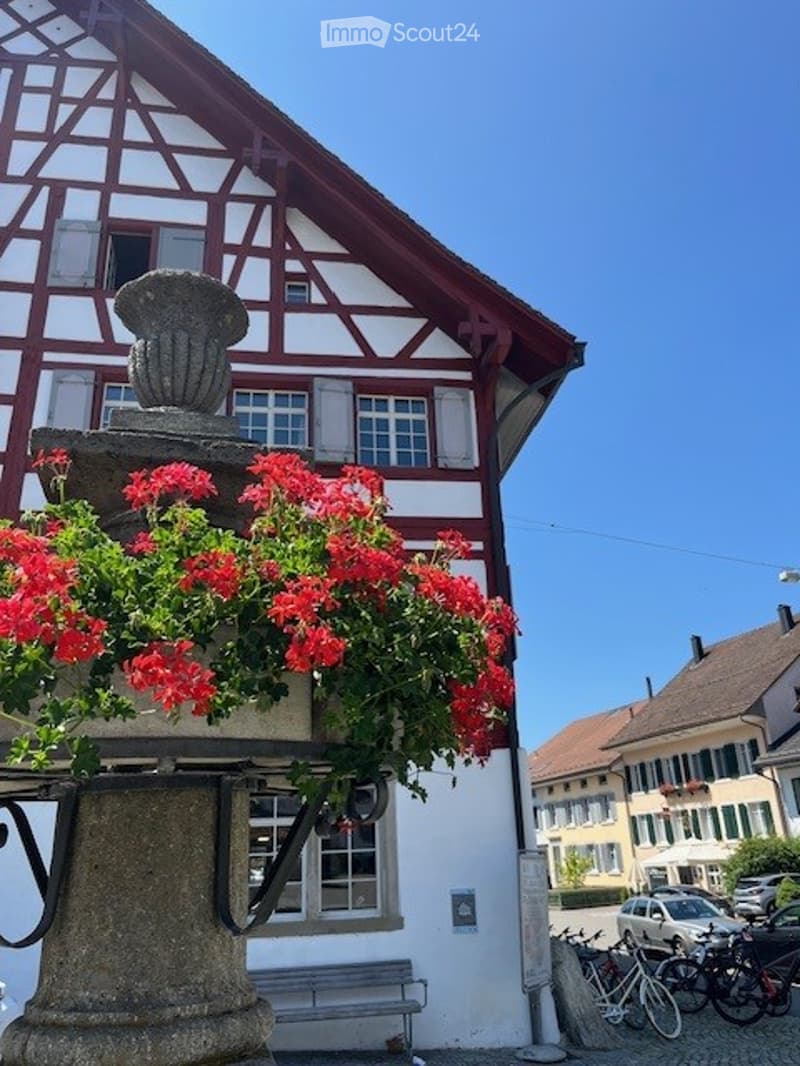 Traditional half-timbered building with red and white facade, multiple windows, and a decorative flower planter with red flowers on the balcony.