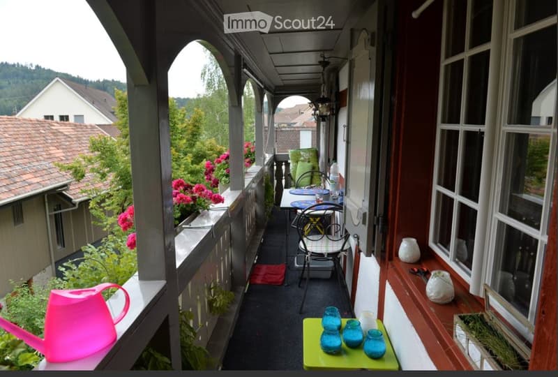 Balcony with a small table and chairs, decorated with vases, a watering can, and colorful cups on a green tray. The balcony has a view of a garden and neighboring houses.